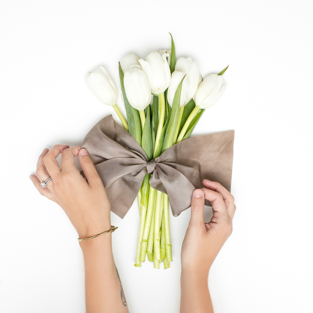 A woman's hands tying a taupe bow around a bouquet of white tulips